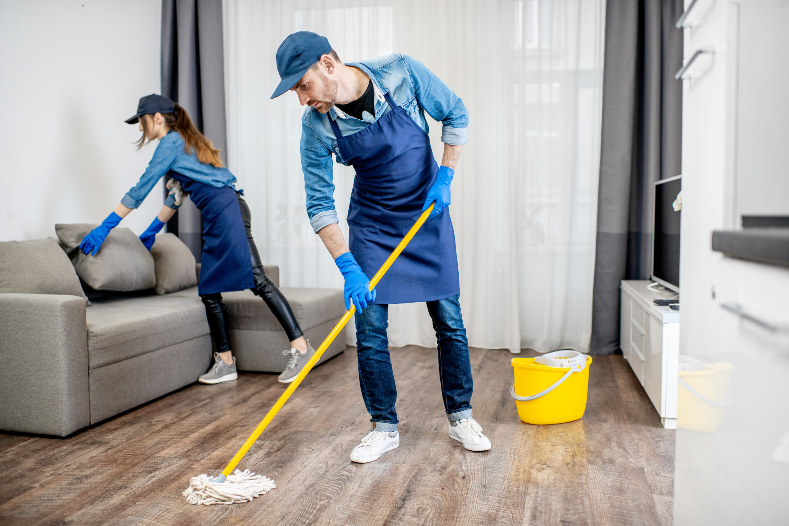 Professional cleaners in blue uniform washing floor and wiping dust from the furniture in the living room of the apartment. Cleaning service concept